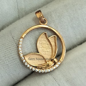 Könnte beinhalten: Goldener Anhänger mit einem Schmetterlingsdesign und einem Kreis aus klaren Steinen am Rand. Der Anhänger liegt auf einer hellbraunen Oberfläche. "Gem house studio" ist auf dem Schmetterling geschrieben.