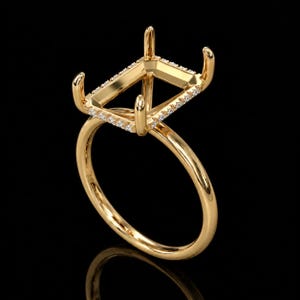 May include: A yellow gold ring setting featuring a rectangular head with four prongs designed to hold a gemstone. The rectangular frame is accented with a row of small, clear pavé-set stones along the top edges of the setting.