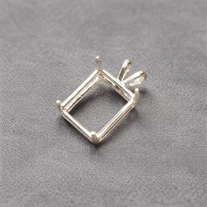 Puede incluir: Un engaste de joyería rectangular plateado con cuatro garras y un lazo para una cadena. El engaste está diseñado para sostener una piedra preciosa rectangular. El engaste está hecho de metal brillante sobre una superficie gris.