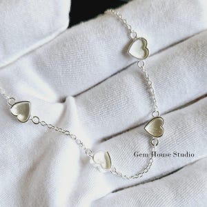 May include: Silver chain necklace with five open heart charms. The chain is delicate and the hearts are small. The necklace is on a white fabric background.