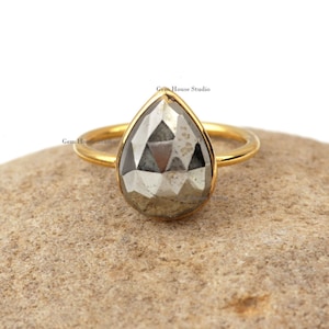 Pode incluir: Um anel de ouro com uma gema de pirita em forma de pêra. O anel está colocado em uma rocha marrom.