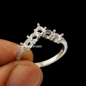 Puede incluir: Un anillo plateado con cinco engastes para piedras preciosas. El anillo tiene un diseño curvo, con los engastes dispuestos en un ligero arco. El texto "GEM HOUSE STUDIO" es visible en el anillo. El anillo está sostenido por una mano sobre un fondo negro.