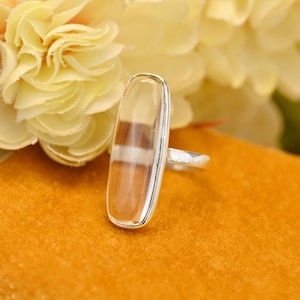 Op de afbeelding: Een zilveren ring met een rechthoekige, heldere edelsteen met bruine en witte banden. De ring is geplaatst tegen een geel-oranje oppervlak, met een vage bloemenachtergrond.