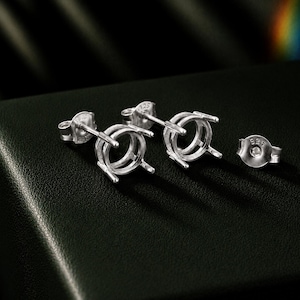 May include: A pair of silver stud earring settings featuring a four-prong basket design for holding round gemstones. Each earring post is stamped with 925, and the set includes two matching silver earring backs, also clearly marked with 925.