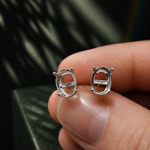 May include: Two silver-colored oval earring settings. Each setting has four prongs and a central bar. The earrings are held by a person's fingers against a dark background.