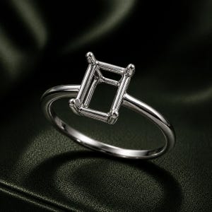 May include: A silver ring setting with a rectangular center, designed to hold a gemstone. The ring has a polished finish and a simple band. The setting features four prongs at each corner to secure the stone.