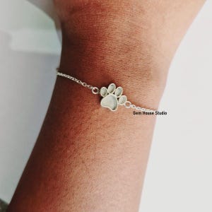 Könnte beinhalten: Ein silbernes Kettenarmband mit einem Pfotenanhänger. Der Anhänger ist silber mit einer weißen Mitte. Das Armband befindet sich am Handgelenk einer Person. Gem House Studio ist auf dem Bild gedruckt.