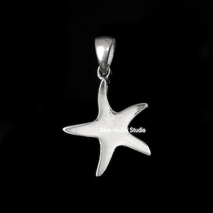 Pode incluir: Um pingente de estrela do mar prateado com uma argola para corrente. O pingente tem formato de estrela de cinco pontas e está sobre um fundo preto. O texto "Gem House Studio" é visível no pingente.