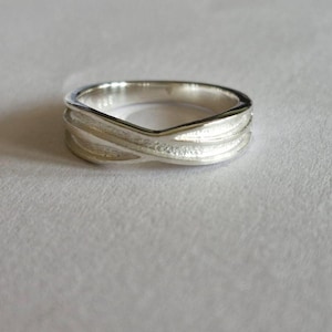 May include: A silver ring with a unique, intertwined design. The ring features a V-shaped top and textured bands that cross over each other, creating an elegant and modern look. The ring is set against a plain, light background.