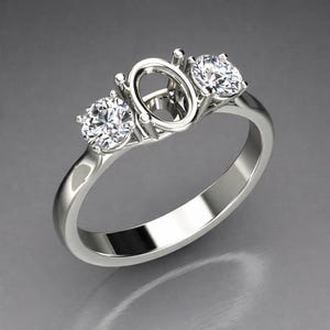 Puede incluir: Un engaste de anillo de compromiso plateado con un centro ovalado y dos piedras laterales redondas. El anillo está sobre un fondo gris, mostrando su metal pulido y piedras brillantes. El anillo está diseñado para sostener piedras preciosas.