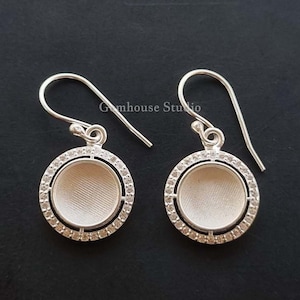 May include: A pair of silver earrings with a round, white, cabochon-style setting surrounded by a circle of clear stones. The earrings have a simple, elegant design.
