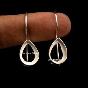 May include: A pair of silver teardrop-shaped earring blanks with a small hole at the top for attaching a hook or finding. The earring blanks are shown on a finger.
