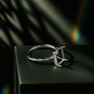 May include: A silver ring setting with a twisted band and a square-shaped head. The ring is displayed on a dark green surface, with shadows and a hint of a rainbow in the background. The ring is designed to hold a gemstone.