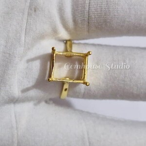 May include: A gold ring featuring a square setting. The ring's design is delicate, with small, rounded details at each corner of the setting. The text "Gemhouse Studio" is visible in the background.