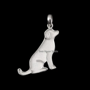 Op de afbeelding: Een zilveren hanger in de vorm van een zittende hond. De hanger heeft een lus voor een ketting. De tekst "Gem House Studio" is zichtbaar op de hanger. De hond kijkt naar rechts.