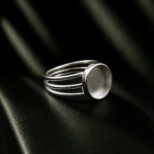 May include: A silver-toned ring featuring a multi-band design with three parallel wires that converge into a single circular bezel setting. The bezel is empty and designed to hold a stone or cabochon, showcasing a polished metallic finish.