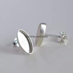 May include: Two silver oval stud earring settings. The settings have a shallow dish shape to hold stones or other materials. The posts and backs are also silver. The earrings are displayed on a light gray background.