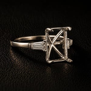 May include: A silver-toned metal ring setting featuring a rectangular prong head designed to hold a center stone. The band is accented on each side by a single tapered baguette-cut clear stone, creating a three-stone style mounting for jewelry.