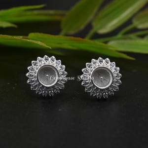 May include: Two silver-colored stud earrings. Each earring features a central recessed area surrounded by a ring of small, clear, petal-shaped accents. The earrings are set against a dark background with green foliage.