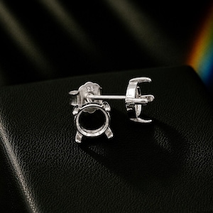 May include: A pair of silver stud earring settings featuring a four-prong basket design for holding a gemstone. The earring backs are clearly stamped with the number 925, indicating sterling silver composition. The metal has a polished finish.