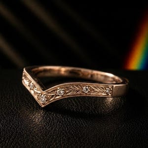 May include: A V-shaped rose gold ring featuring an engraved leaf pattern along the band and five small, round, clear stones set at intervals. The metal has a polished finish with milgrain detailing along the edges of the band.