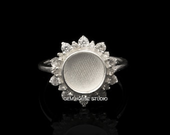 Op de afbeelding: Een zilveren ring met een ronde, witte stenen zetting omgeven door kleinere, heldere stenen. De ring ligt op een zwarte achtergrond. GEM HOUSE STUDIO is onder de ring gedrukt.
