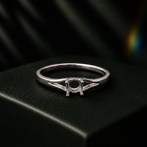 May include: A silver ring with a round setting and split shank design. The ring is set against a dark background, highlighting its polished metal finish. The ring is designed to hold a gemstone.