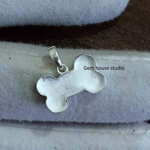 May include: Silver dog bone charm pendant with a smooth, polished finish. The pendant is shaped like a bone and has a loop at the top for attaching to a chain or bracelet. Gem house studio is printed on the pendant.