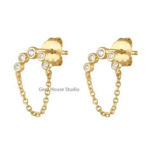 May include: A pair of gold-colored stud earrings with a chain. Each earring features a curved row of small, clear gemstones and a delicate chain that dangles below. The earrings are set against a white background.