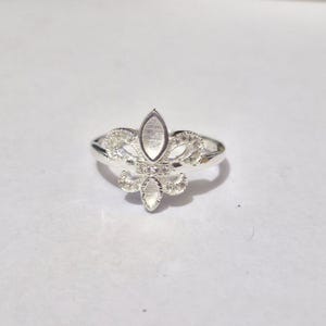 May include: A silver ring with a fleur-de-lis design. The ring features a central oval-shaped stone and smaller stones, with intricate filigree detailing. The ring is set against a white background, highlighting its elegant design.