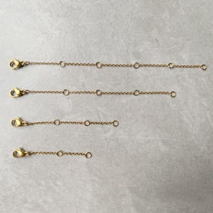 May include: Four gold-colored chain extenders with lobster claw clasps and small circular loops. The extenders vary in length, with the longest at the top and the shortest at the bottom. The chains are displayed on a light gray surface.