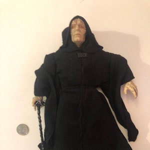 May include: A dark gray hooded robe with a long, flowing design. The robe is worn by a figure with a pale, wrinkled face and a long, pointed nose. The figure is holding a black staff in their right hand.