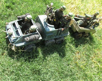 May include: A green military toy vehicle with a white skull and horns on the front. The vehicle has a crew of four toy soldiers wearing green camouflage uniforms and helmets. The vehicle is on a grassy surface.