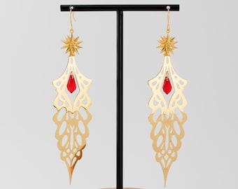 May include: Gold filigree earrings with red crystal accents. The earrings are displayed on a wooden stand.