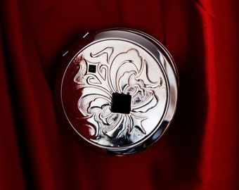 May include: A silver compact mirror with an ornate floral design. The mirror has a small square opening in the center.