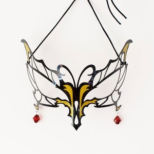 Può includere: Maschera da ballo in maschera nera e oro con un design decorativo. La maschera ha una forma simmetrica con intricati motivi ritagliati e due accenti di gioielli rossi appesi dal basso. Un cordino nero è attaccato per indossarla.