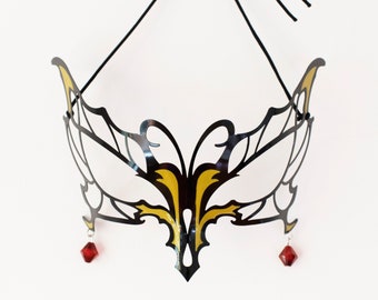 May include: Black and gold masquerade mask with a decorative design. The mask has a symmetrical shape with intricate cut-out patterns and two red jewel accents hanging from the bottom. A black string is attached for wearing.