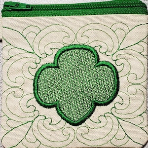 Może przedstawiać: Mała, biała kosmetyczka z zamkiem błyskawicznym z zielonym haftowanym logo Girl Scout w środku. Logo jest otoczone ozdobnym, haftowanym wzorem.