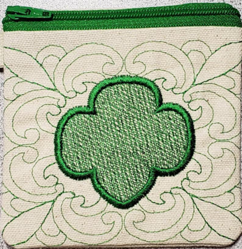 Może przedstawiać: Mała, biała kosmetyczka z zamkiem błyskawicznym z zielonym haftowanym logo Girl Scout w środku. Logo jest otoczone ozdobnym, haftowanym wzorem.