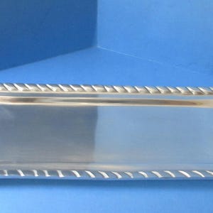 May include: A rectangular silver-colored serving tray with rope-like detailing around the edges and at the handles. The tray has a smooth, reflective surface and is designed with nautical-themed accents.