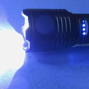 May include: A black handheld flashlight with a crenelated bezel emitting a bright white beam. The device features a circular power button illuminated with a blue ring and a vertical row of four small blue LED indicator lights on the side.