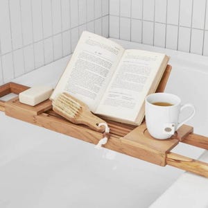 May include: A wooden bathtub tray holds an open book, a white mug with tea, a bar of soap, and a wooden brush. The tray has a natural wood finish and is designed to rest across the bathtub.