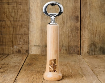 May include: A wooden bottle opener with a silver top and a Seattle Mariners logo. The opener is standing on a wooden surface.