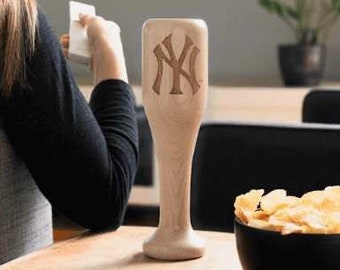 May include: A wooden baseball bat-shaped beer glass with the New York Yankees logo carved into it. The glass is sitting on a table in front of a person watching a baseball game on TV.