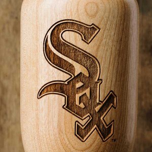 Puede incluir: Un bate de b&eacute;isbol de madera con un logotipo "S" estilizado de color marr&oacute;n oscuro quemado en la superficie. El logotipo es una marca registrada de los Chicago White Sox.