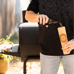 May include: A person pouring a dark liquid from a bottle into a wooden baseball bat-shaped cup with a compass design and the text "Little League" engraved on it.