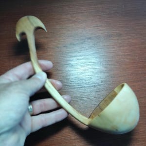May include: A wooden ladle with a curved handle and a rounded bowl. The handle has an arched design at the top. The ladle is made of light-coloured wood. The item is new and appears to be handmade.