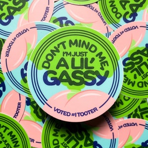 May include: A close-up of several round stickers with a green, blue, and pink design. The stickers feature the text "Don't Mind Me, I'm Just A Lil' Gassy" in a bold, colorful font. The stickers are printed with the hashtag "#ITOOTER" and the words "Voted #1".