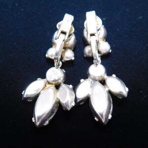Op de afbeelding: Een paar zilverkleurige clip-on oorbellen met heldere strass-accenten. De oorbellen hebben een cluster van strass in het midden en een hangende cluster van strass.