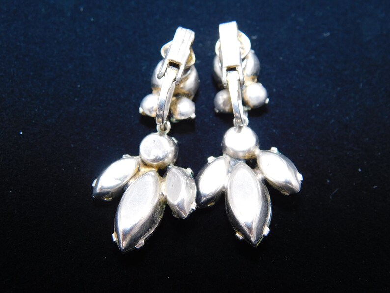 Op de afbeelding: Een paar zilverkleurige clip-on oorbellen met heldere strass-accenten. De oorbellen hebben een cluster van strass in het midden en een hangende cluster van strass.